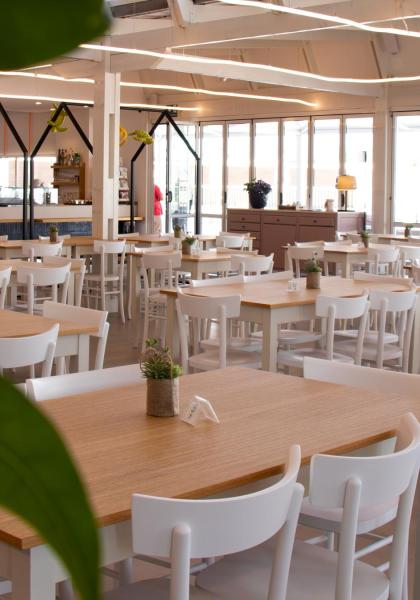Ampia sala luminosa con tavoli in legno e sedie bianche, atmosfera accogliente.