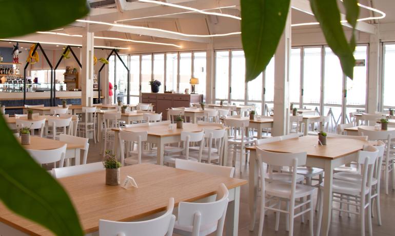 Ampia sala luminosa con tavoli in legno e sedie bianche, atmosfera accogliente.