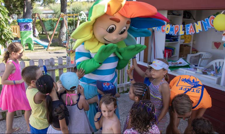 Bambini felici giocano con una mascotte colorata in un parco giochi soleggiato.