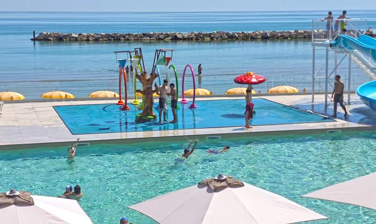 Piscina colorata con giochi d'acqua, scivoli e mare sullo sfondo.