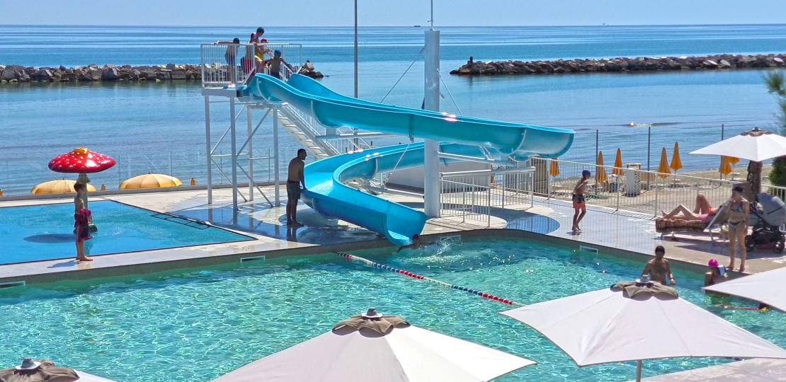 Piscina soleggiata con scivolo blu, mare calmo e relax estivo.
