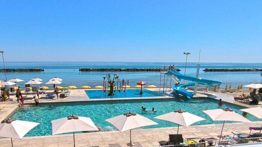 Piscina soleggiata con scivolo, ombrelloni e mare calmo in sottofondo.