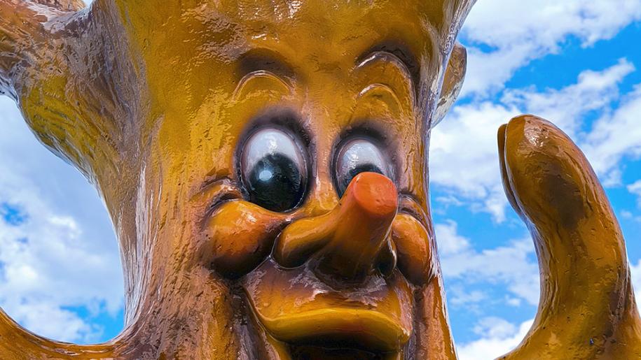 Statua di albero sorridente, occhi grandi, cielo azzurro, atmosfera giocosa e allegra.